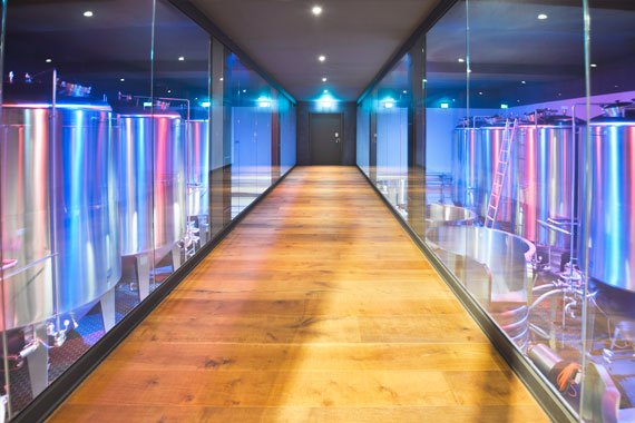 Ein Gang mit Holzboden, seitlichen Glasfronten mit Blick auf die Braukessel, modern beleuchtet.